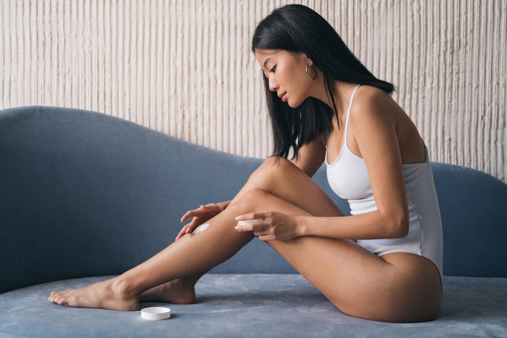 Korean woman applying body cream on her legs, sitting on couch at home