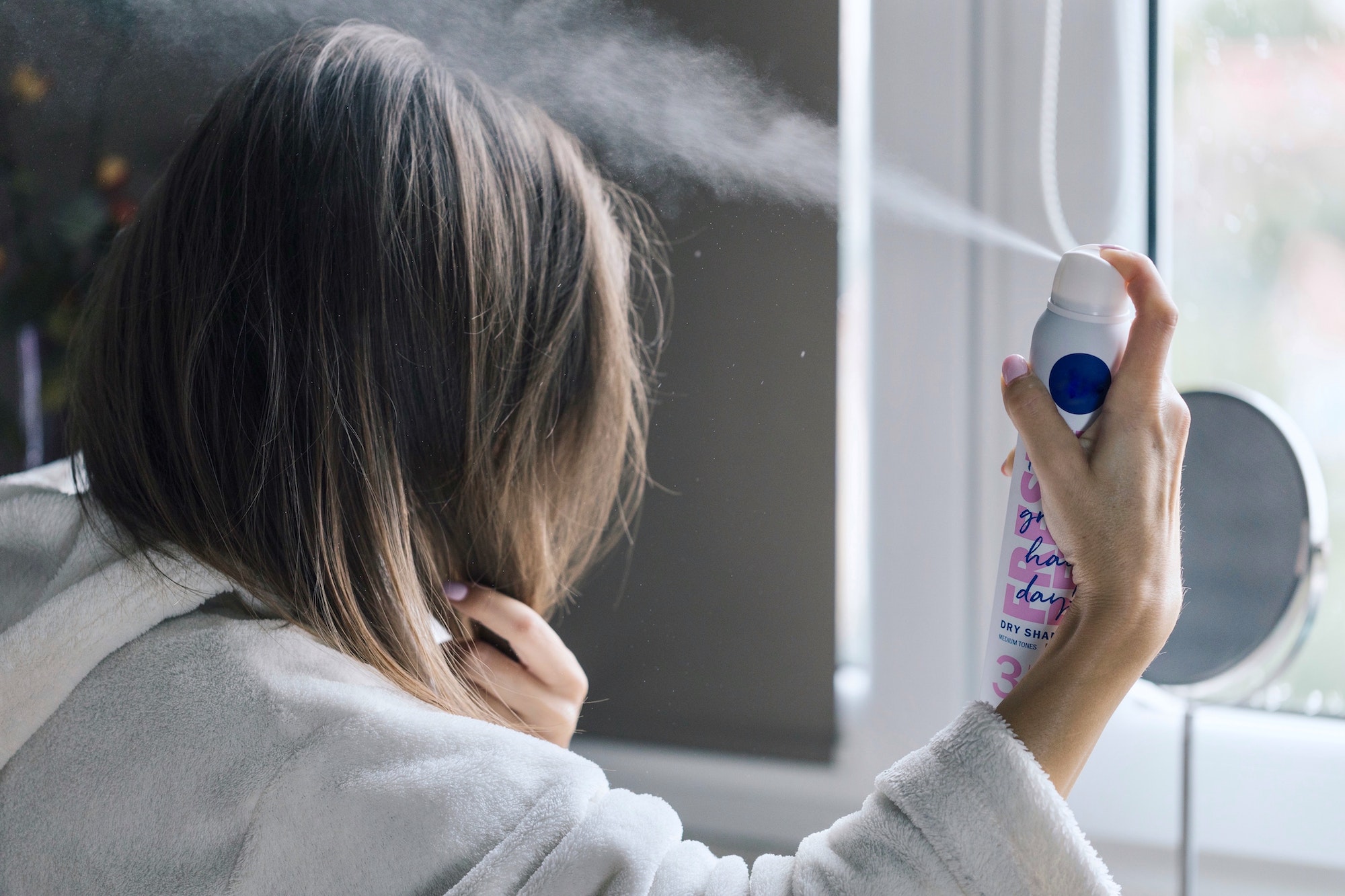 En dame med langt, brunt hår sprayer tørrsjampo i røttene mens hun står foran speilet på badet. Hun masserer produktet inn i håret med fingrene og børster deretter gjennom håret med en kam for å fjerne overflødig produkt. Håret hennes ser nå renere og friskere ut, som om hun nettopp har vasket det.