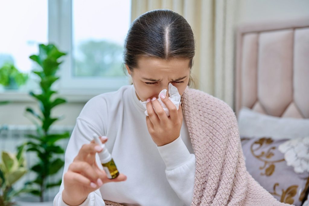 Teenage girl with runny nose treating rhinitis using nose drops spray