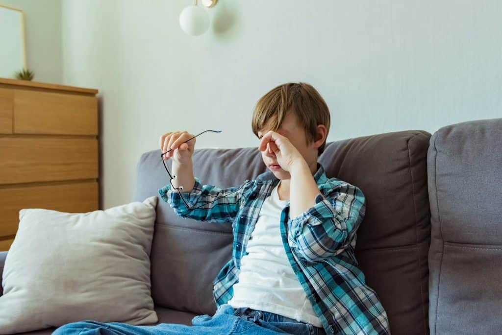Small school boy rubs his eyes from using glasses for myopia at home. Dry eyes. Pain. Bad eyesight.